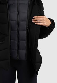 Jachetă puffer neagră cu fermoar frontal, având un căptușeală matlasată. Materialul exterior este neted, cu un finisaj mat.