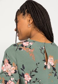 Blusa floral verde de mangas curtas, com padrões de flores em rosa e preto, uma pequena abertura nas costas e uma textura de tecido suave.