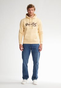 Felpa gialla con logo "Petrol Ind Co", tasca a marsupio, cordini regolabili, abbinata a jeans blu e sneakers bianche.
