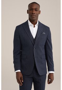 Costume bleu marine à rayures avec une coupe ajustée, veste croisée à un bouton et revers en pointe. Chemise blanche à manches visibles, détail de pochette.