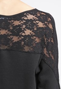 Haut noir comportant un panneau en dentelle florale sur les épaules, avec un corps en tissu lisse et une coupe décontractée. Encolure arrondie et manches longues.