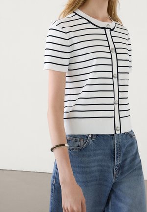 Femme portant un cardigan blanc à manches courtes avec des rayures horizontales noires et des boutons argentés, assorti à un jean en denim bleu et un bracelet rigide.