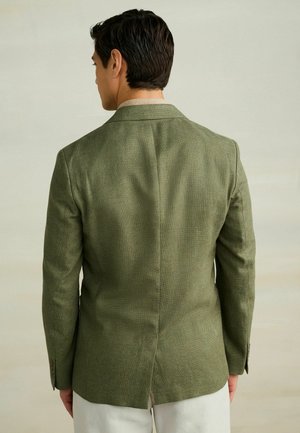 Texturált olívazöld blézert viselő férfi, aki hátat fordít egy semleges háttér előtt, világos színű nadrággal párosítva.