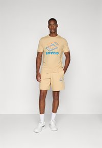 Conjunto de t-shirt bege e calções com um logo e design gráfico em azul e laranja da "Lotto". Feito de material de algodão suave. Ténis brancos usados.
