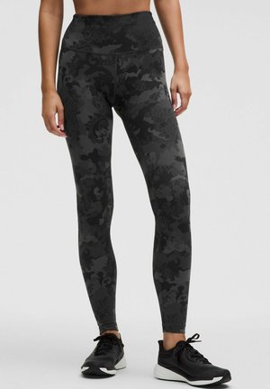 Schwarze Leggings mit einem strukturierten Blumenmuster, hoch geschnittenem Design und einer figurbetonten Silhouette, kombiniert mit schwarzen Sportschuhen.