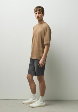 Junger Mann steht, trägt ein braunes übergroßes T-Shirt, dunkelgraue Shorts, weiße Socken und weiße Sneaker vor einer einfarbigen hellen Wand.