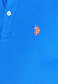 Modra polo majica iz teksturirane kotna, ki ima tri gumbe in oranžni vezeni logotip na levem prsnem delu.