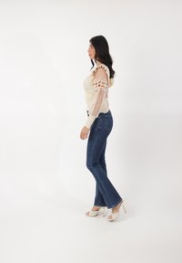 Top beige con maniche trasparenti e arricciate con dettagli in pizzo, abbinato a jeans blu scuri a zampa d'elefante e sandali aperti color crema.