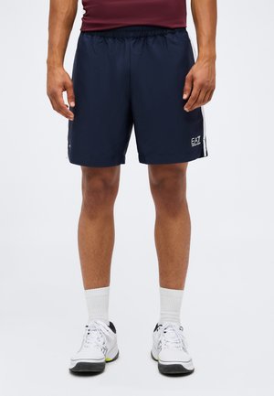 Mann trägt dunkelblaue EA7-Shorts, weiße Sportsocken und weiße Sneaker, steht vor einem schlichten hellen Hintergrund.