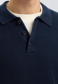 Primer plano de una persona que lleva una camiseta polo de punto texturizado azul marino sobre una camiseta blanca de cuello redondo.