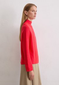 Roter Pullover mit hohem Kragen aus Strickmaterial, mit langen Ärmeln und lässigem Schnitt, kombiniert mit einem beigen Rock.