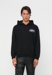 Sweatshirt noir avec capuche, poche frontale et un graphique coloré sur le côté gauche de la poitrine. Le tissu paraît doux avec une coupe décontractée.