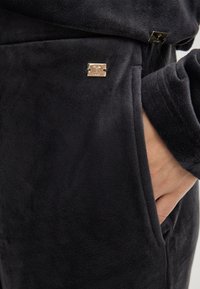 Zwarte fleece loungewear met een zachte textuur, zijzak en een kleine metalen logoaccent near het taillegebied.