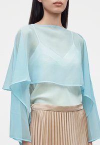 Femme portant un haut transparent bleu clair à manches longues par-dessus un caraco blanc, associé à une jupe plissée beige.