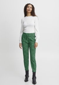 Weißes, geripptes Langarmshirt mit gepufften Schultern, kombiniert mit grünen gemusterten Hosen mit geometrischen Designs und schwarzen Knöchelstiefeln.
