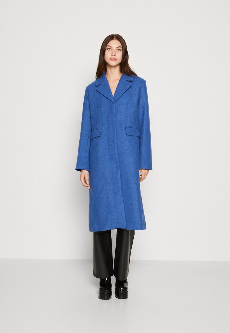 Manteau long en laine bleu avec un col à revers, deux poches avant et une silhouette droite, associé à un pantalon noir et des chaussures noires à semelles épaisses.