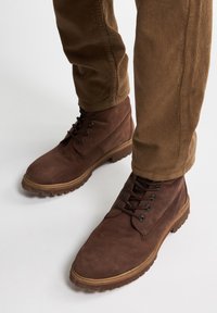 Bruine suède enkelboots met vetersluiting en rubberen zool, gecombineerd met lichtbruine corduroy broek met een geribbelde textuur.