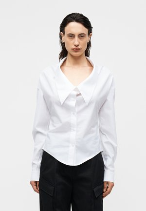 Frau mit dunklen Haaren, die ein weißes Hemd mit breitem Kragen und schwarze Hosen mit Seitentaschen vor einem einfarbigen Hintergrund trägt.