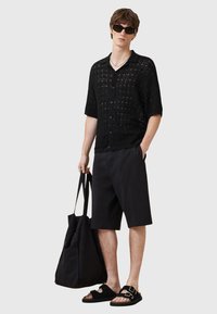 Camiseta negra de punto de manga corta, pantalones cortos negros, sandalias de slide negras y un gran bolso tote negro. Los accesorios incluyen gafas de sol y un collar.