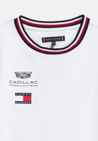 Άσπρο μπλουζάκι με λαιμόκοψη crew neck και κόκκινο, άσπρο και ναυτικό ριγέ γιακ ά, με λογότυπα της ομάδας Cadillac Formula 1 και της Tommy Hilfiger στο στήθος.