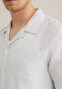Svetlo siva kratka majica s kratkimi rokavi in gumbi, z diskretnim geometričnim vzorcem, žepom na prsih in zasnovo ovratnika kamp stila. Gladka tekstura tkanine.