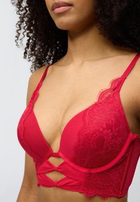 Κόκκινο μπραντ με δαντέλα και ύφασμα, με βαθύ ντεκολτέ, ρυθμιζόμενους ιμάντες και κοψίματα στη ζώνη για επιπλέον υφή και οπτικό ενδιαφέρον.