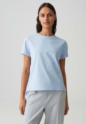 Femme aux longs cheveux foncés portant une chemise bleu clair à manches courtes et un pantalon gris clair sur un fond clair uni.
