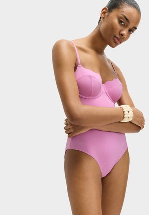 Kvinde med armene over kors iført en lyserød, tætsiddende bodysuit og chunky cremefarvet armbånd mod en ensfarvet lys baggrund.