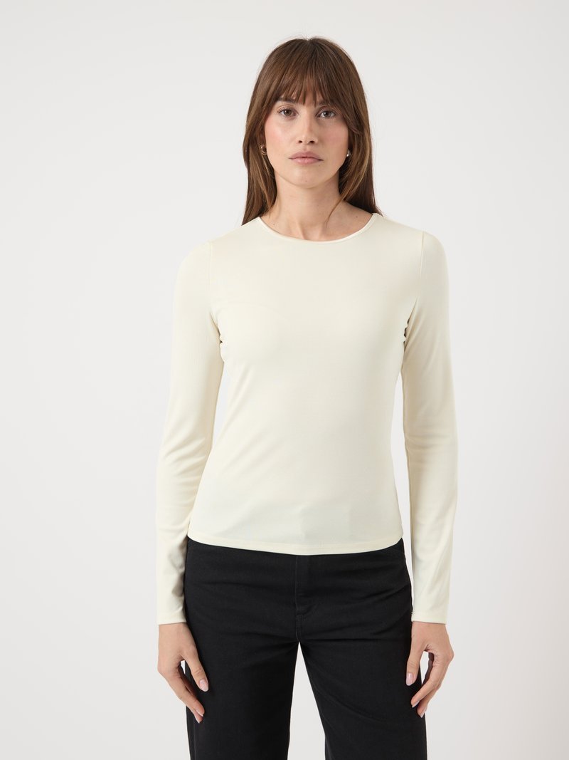 Vrouw met recht bruin haar, gekleed in een crèmekleurige lange mouwtop en zwarte broek, staande tegen een effen witte achtergrond.