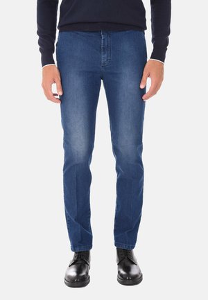 Uomo con jeans slim fit blu, scarpe stringate in pelle nere e una maglia scura a maniche lunghe, in piedi con le braccia rilassate ai lati.