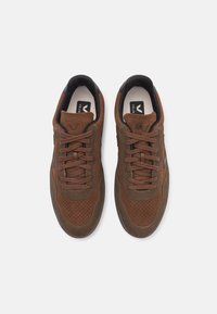 Bruna mockaskor med perforeringar, platta snörningar och en svart detalj på hälen, med en logotyp på tungan.