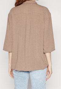 Donna che indossa una camicia marrone ampia e testurizzata con maniche a tre quarti e jeans blu chiaro, vista di spalle su uno sfondo bianco semplice.