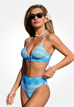 Kobieta w niebieskim bikini z wzorem, z wysokim stanem, naszyjnikiem z muszli, bransoletką, okularami przeciwsłonecznymi i kwiatem we włosach na białym tle.