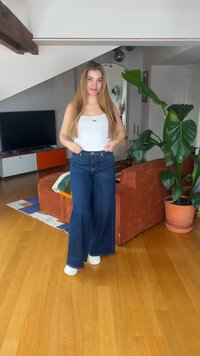 Young woman with long hair wearing white top and wide-leg jeans standing in modern living room with orange sofa and large plant.