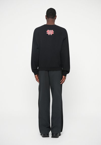 Mand iført sort sweatshirt med rødt og hvidt logo på øvre ryg, løse sorte bukser og sorte sko, står vendt mod en hvid væg.