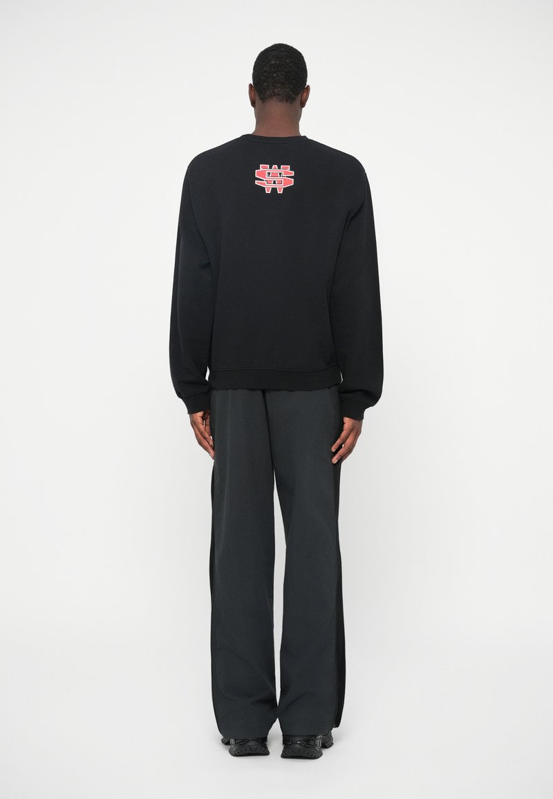 Man in een zwart sweatshirt met een rood en wit logo op de bovenrug, losse zwarte broek en zwarte schoenen, staand met de rug naar een witte muur.