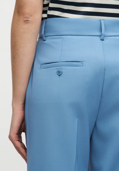 Κοντινό πλάνο ατόμου που φοράει ανοιχτό μπλε παντελόνι με ραμμένη τσέπη στο πίσω μέρος με κουμπί και ριγέ μπλούζα, το χέρι του χαλαρό δίπλα στο σώμα.