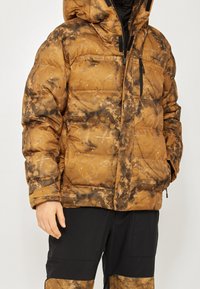 Braune Pufferjacke mit einem strukturierten, marmorierten Muster. Verfügt über eine Kapuze, einen Frontreißverschluss und mehrere Taschen. Kombiniert mit schwarzen Hosen.