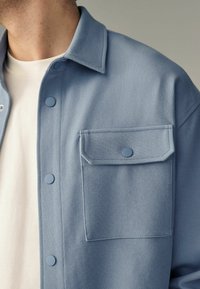 Camisa de botón azul claro con un patrón texturizado, que presenta un bolsillo en el pecho y botones azules redondos. Llevada sobre una prenda blanca.