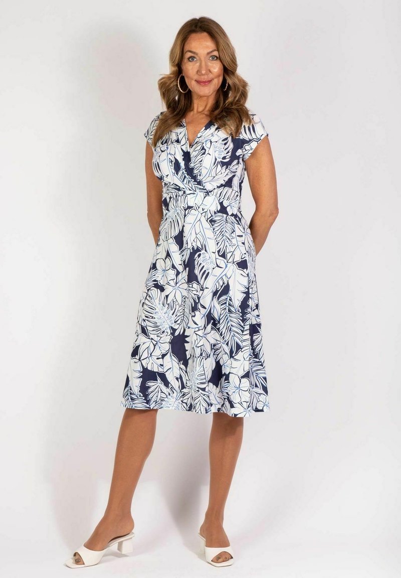 Marineblaues Kleid mit weißem Blumenmuster, kurzen Ärmeln, V-Ausschnitt und ausgestellt Rock. Modell trägt weiße Absatzsandalen.