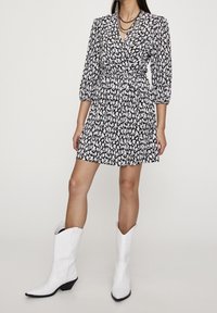 Robe portefeuille à motif floral en noir et blanc, avec des manches trois-quarts, une taille cintrée et une jupette évasée, associée à des bottes de cowboy blanches.
