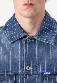 Farmerdzseki, kék csíkozott mintázattal, kiemelkedő gallérral, egyetlen patenttal záródik, logó címke a bal oldalon, enyhén megkopott textúrával.