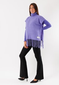 Poncho viola a collo alto con orlo frangiato, abbinato a pantaloni neri aderenti a zampa d'elefante e tacchi a punta neri. Texture morbida, design casual.