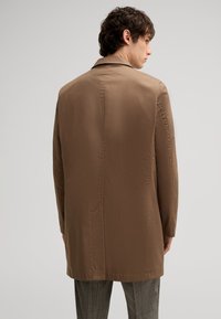 Brauner, leichter Mantel mit glatter Textur, geradem Schnitt und minimalen Nähdetails, getragen über karierter Hose.