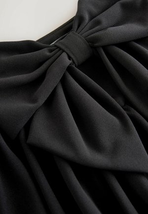 Black fabric dress detail featuring a bow at the waist, with soft texture and pleated design creating folds around the garment.