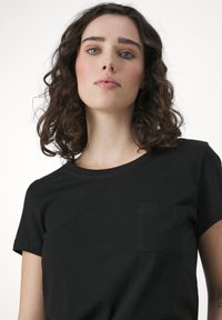 Fekete pamut póló rövid ujjakkal, kerek nyakkivágással és egy kis mellzsebbel. Sima textúrájú és egyszerű dizájnú.