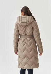Beige-padded takki, jossa on haarukkavalmisteinen kuvio, huppu ja vyö vyötäröllä. Pitkät hihat ja polvenpituinen malli tarjoavat lämpöä ja peittävyyttä.