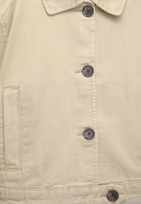 Giacca in tela beige con colletto a camicia, sei bottoni grigi e una tasca laterale, con dettagli di cucitura in vista e una superficie testurizzata.