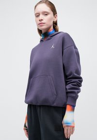 Sudadera morada con bolsillo frontal y logo plateado; combinada con pantalones oscuros que tienen acentos de tie-dye colorido en las mangas.