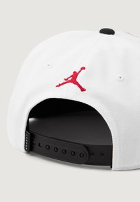 Bjela kapa s crvenim izvezenim Jumpman logotipom, ravnim vizirjem i crnom podesivom trakom straga. Tekstuirana tkanina s ventilacijskim rupama.